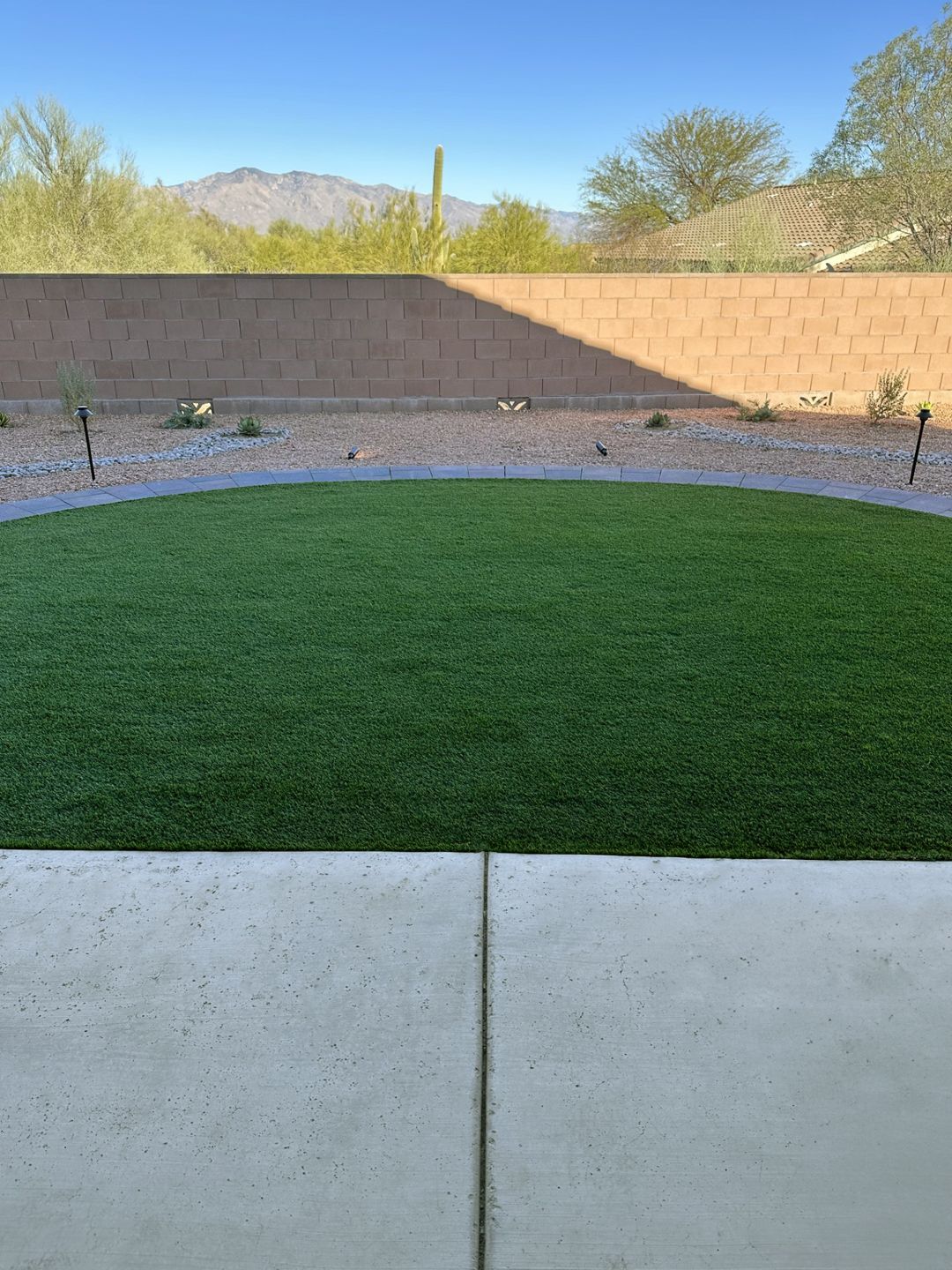 Concrete patio leads to a circular green lawn framed by a tan wall and desert landscape under a blue sky.