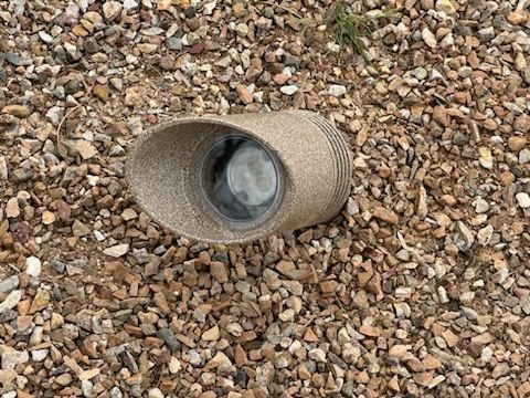 A woven, cylindrical object with a round opening sits on a bed of gravel.