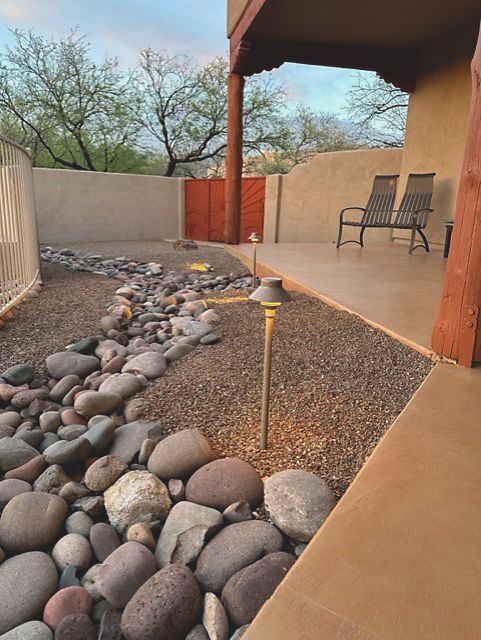 Outdoor space with a rock riverbed, gravel, and pathway lighting. A covered patio features chairs and a rustic gate.