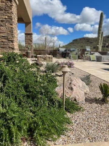 Desert landscape with green shrubs, large rocks, a pathway, and a bright blue sky with clouds.