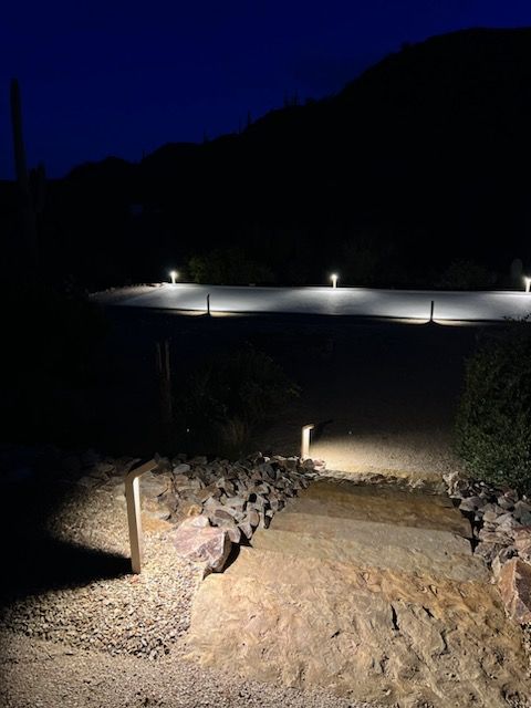 Pathway lit by lamps at night, leading up a stone-lined pathway. Dark blue sky and mountain in the background.