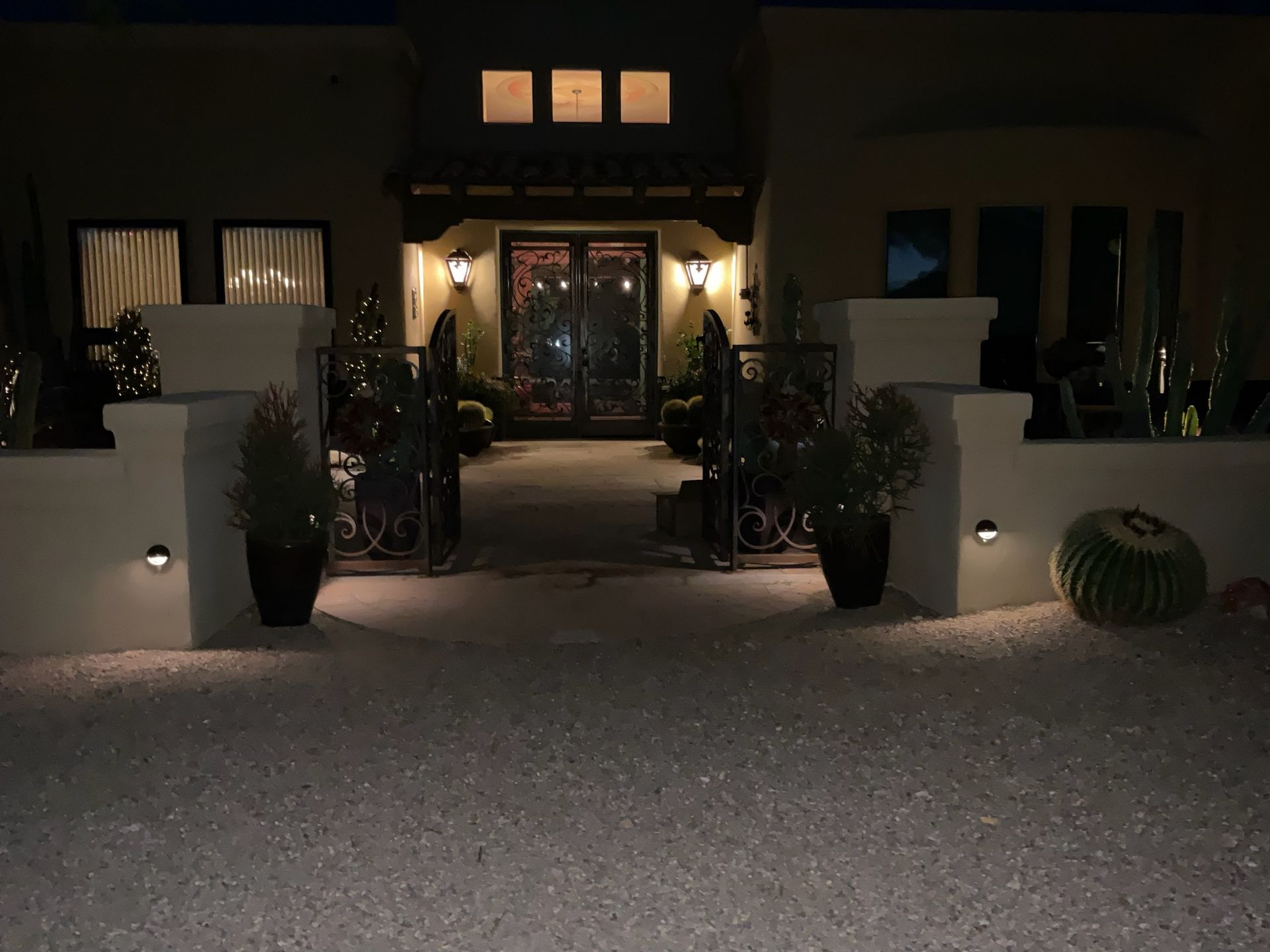 Lit entrance to a home at night. Decorative gate and pillars flank the walkway to the front door, with outdoor lights.