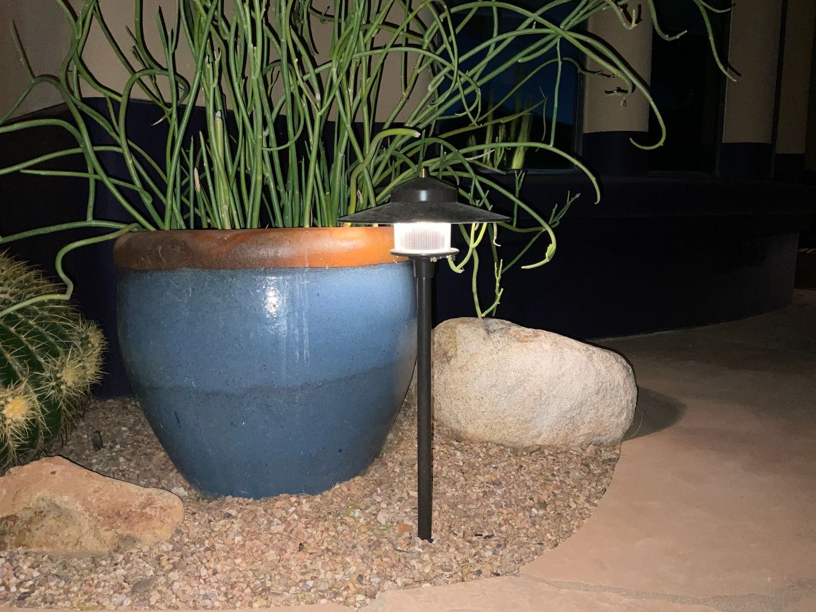 Blue ceramic pot with a plant next to a rock and a black outdoor lamp on a gravel surface.