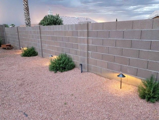 Outdoor scene: A gravel yard with a concrete block wall, illuminated by path lights. Small bushes are planted near the wall.