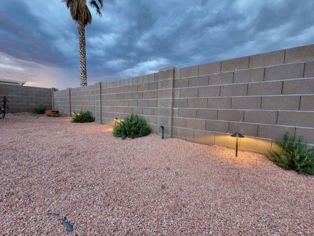 Backyard with gravel ground, block wall, and illuminated bushes under cloudy sky.