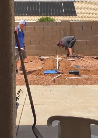Two men working on irrigation in a dirt backyard. One stands, the other is bent over pipes.