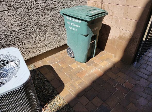 Green trash bin against a wall, on brick pavers, next to an air conditioning unit.
