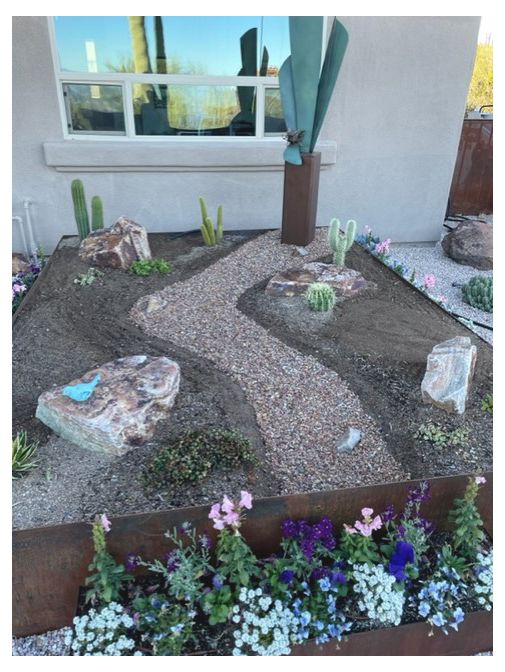 Cactus and rock garden bed with a gravel path, metal sculpture, and blooming flowers along the edge.