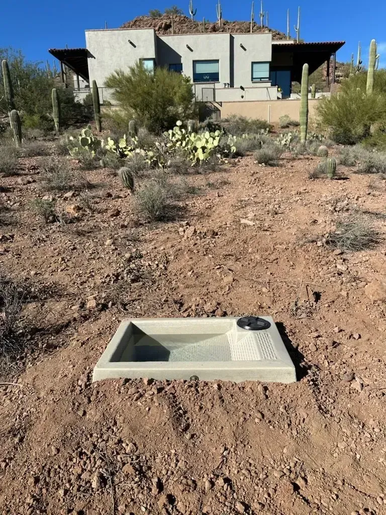 Square water basin in dirt, near a house on a hillside.