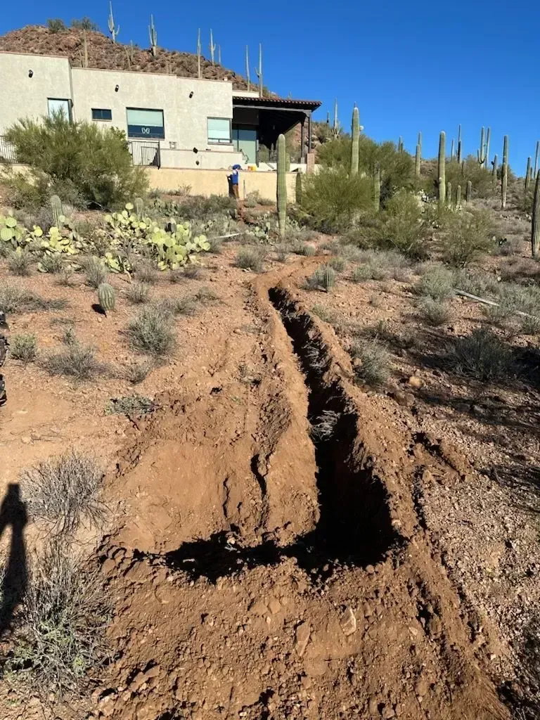 Trench dug in arid, brown terrain with a house and cacti in the background on a sunny day.