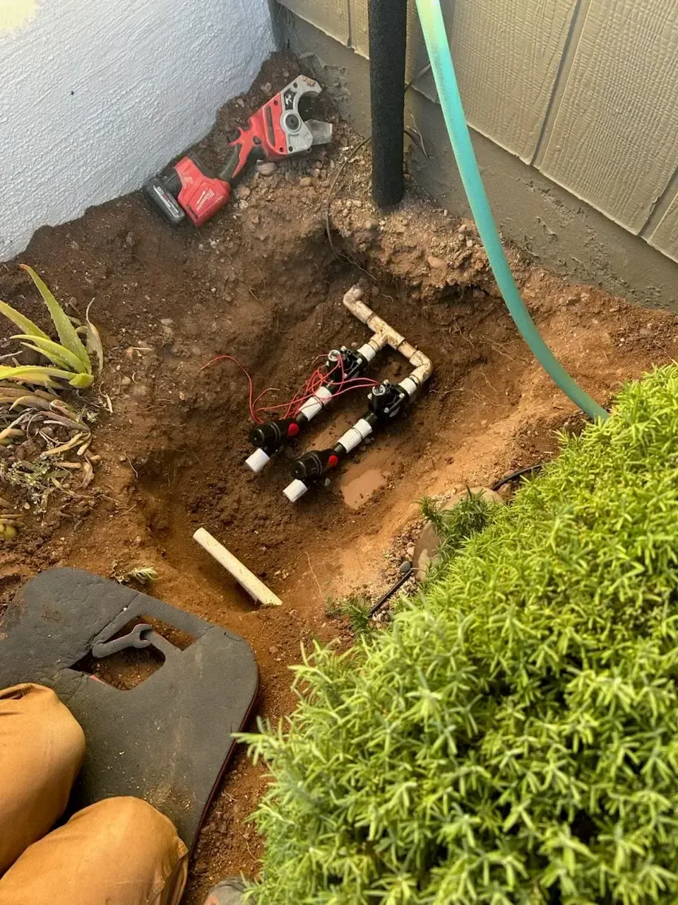 Irrigation system components in an excavated area next to greenery and a wall; wrench and hose present.