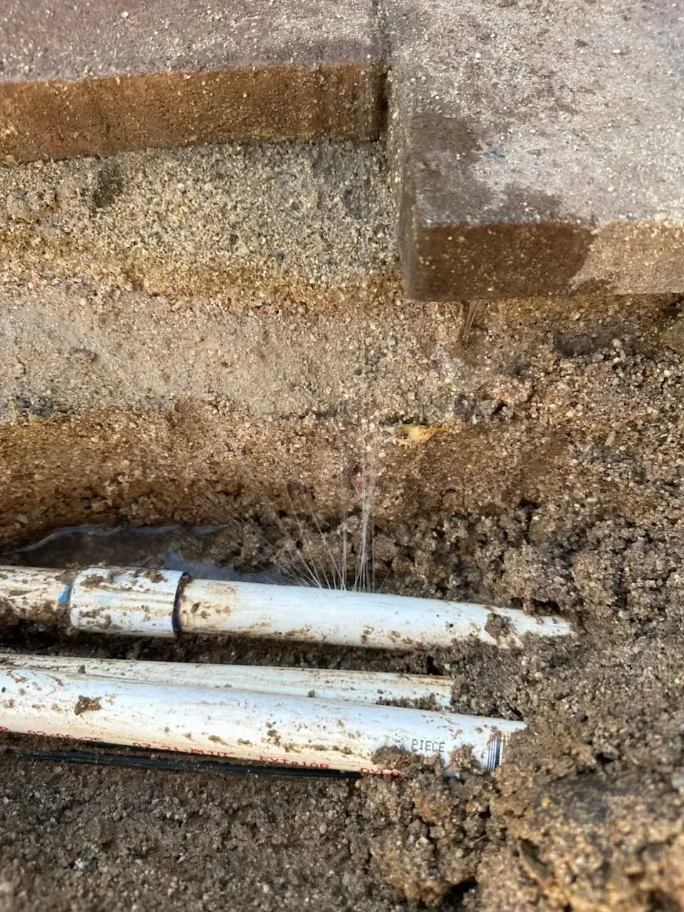 Broken white PVC pipes in a sandy trench, with water spraying upwards. Brick pavers are visible above.
