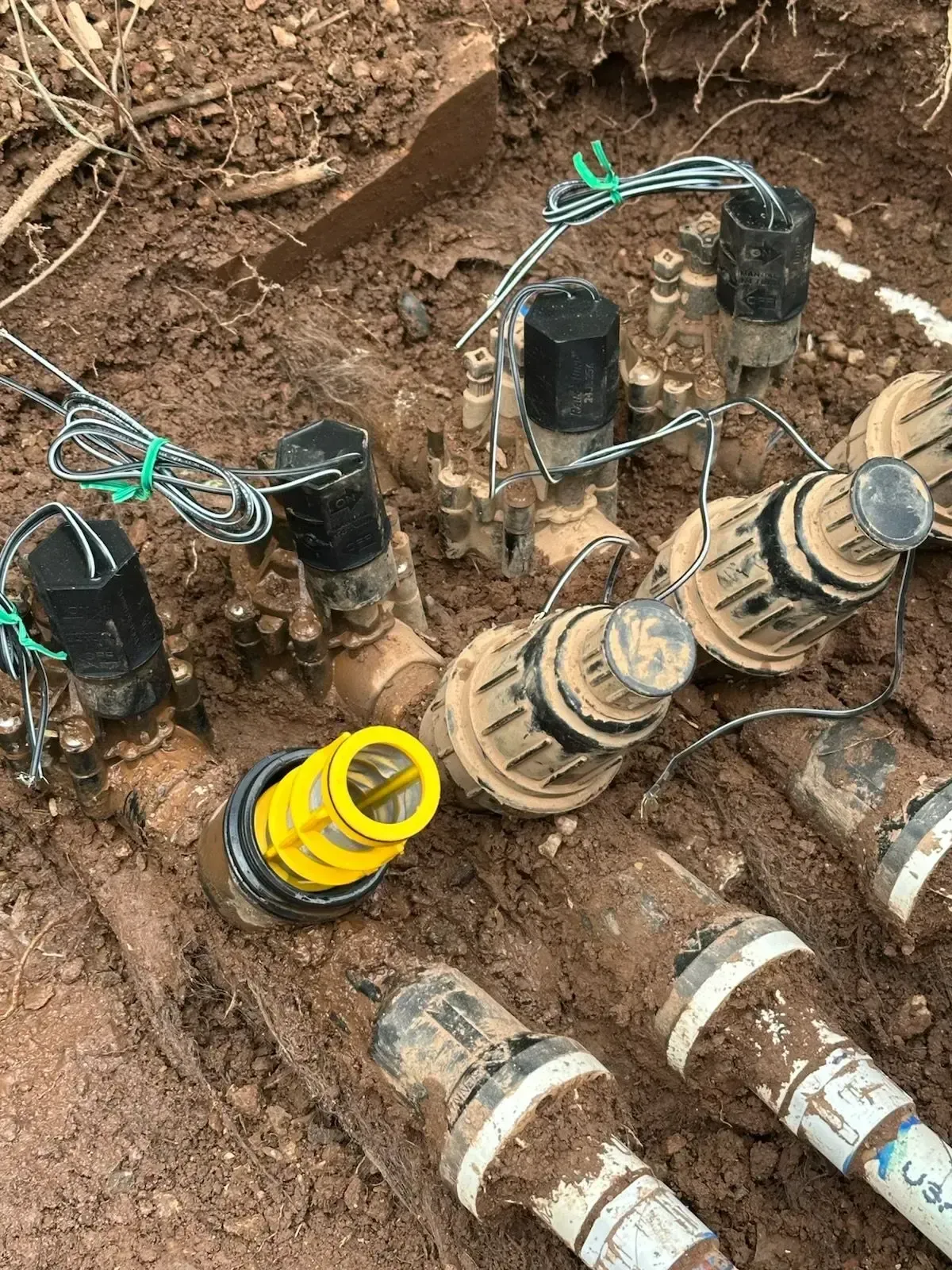 Irrigation system valves and pipes exposed in a muddy trench. Yellow valve cover prominent. Black caps and green wires visible.
