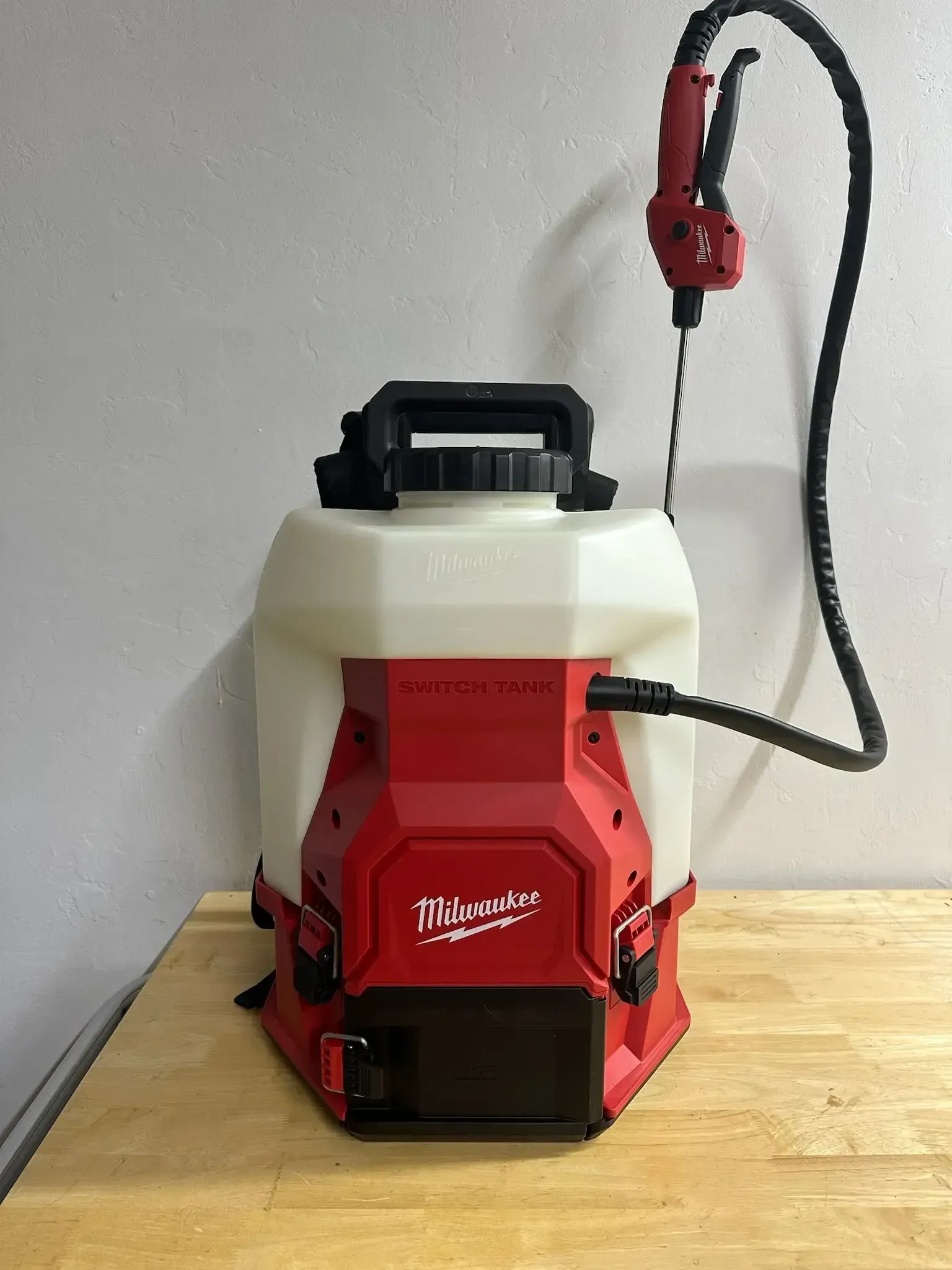 Red and white Milwaukee battery-powered backpack sprayer on a wood table, against a gray wall.