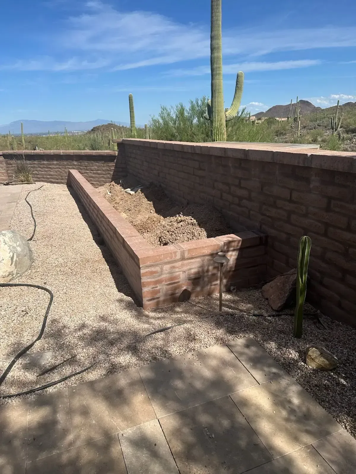 Brick planter box with gravel ground, in front of a desert landscape with cacti and mountains.