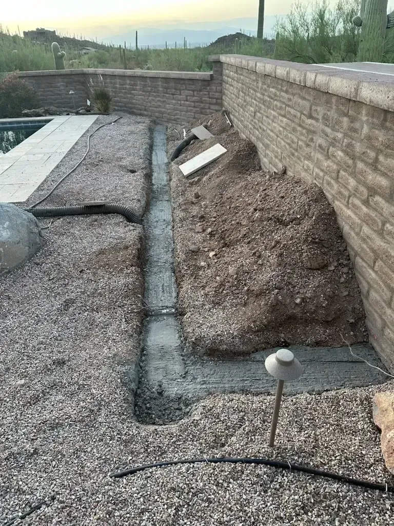 Construction of a concrete drainage channel next to a brick wall and pool. Ground covered in gravel and mulch.
