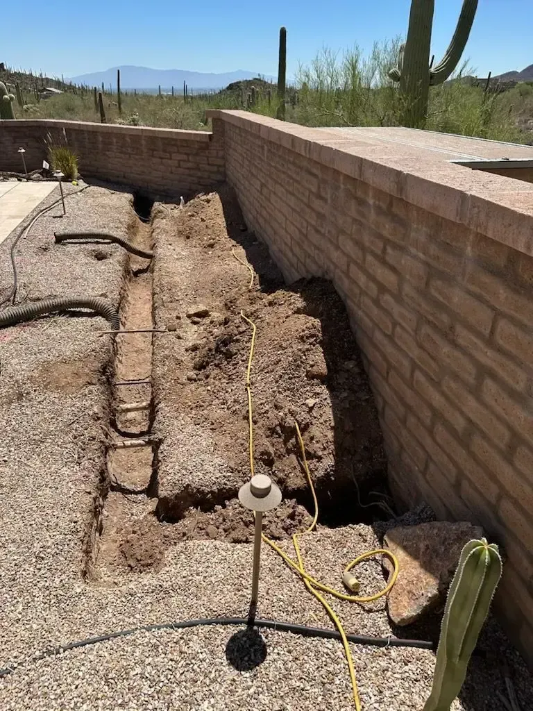 A trench dug along a brick wall in a desert landscape; electrical cord and stakes present.