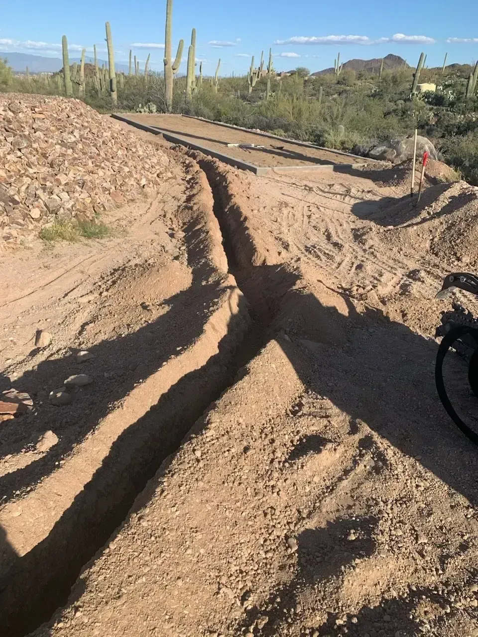 Trench dug in dirt, likely for utility lines, next to road in desert with saguaro cacti.