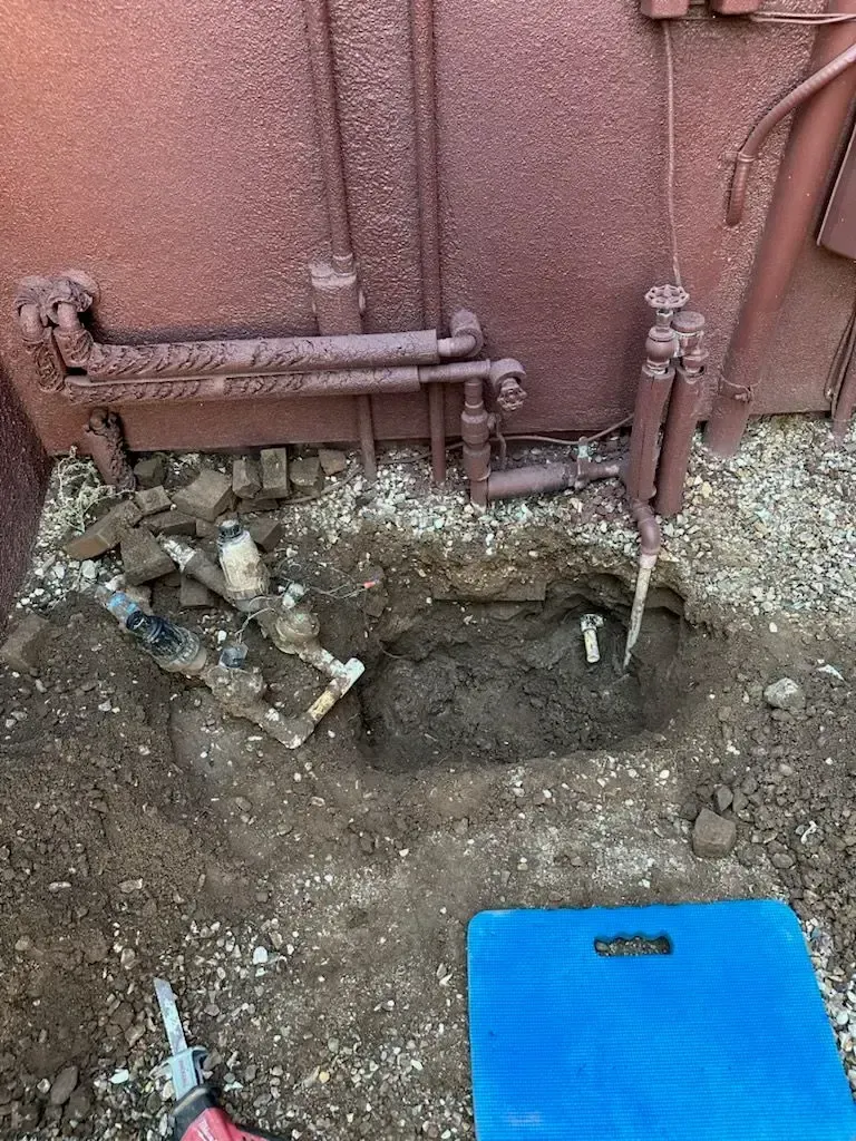 Hole dug near a brown building with exposed pipes, tools, dirt, and a blue kneepad.