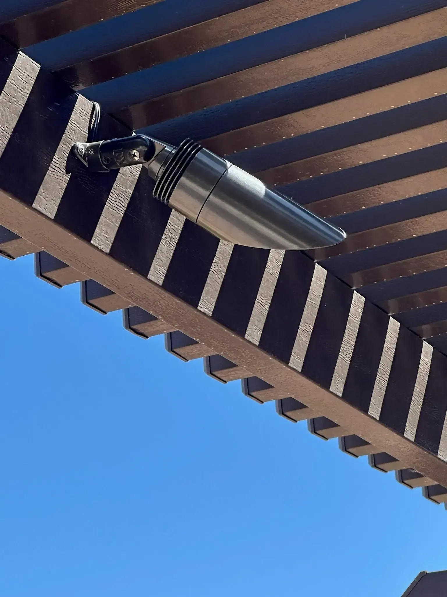 Silver outdoor light fixture mounted on a brown pergola with a blue sky background.