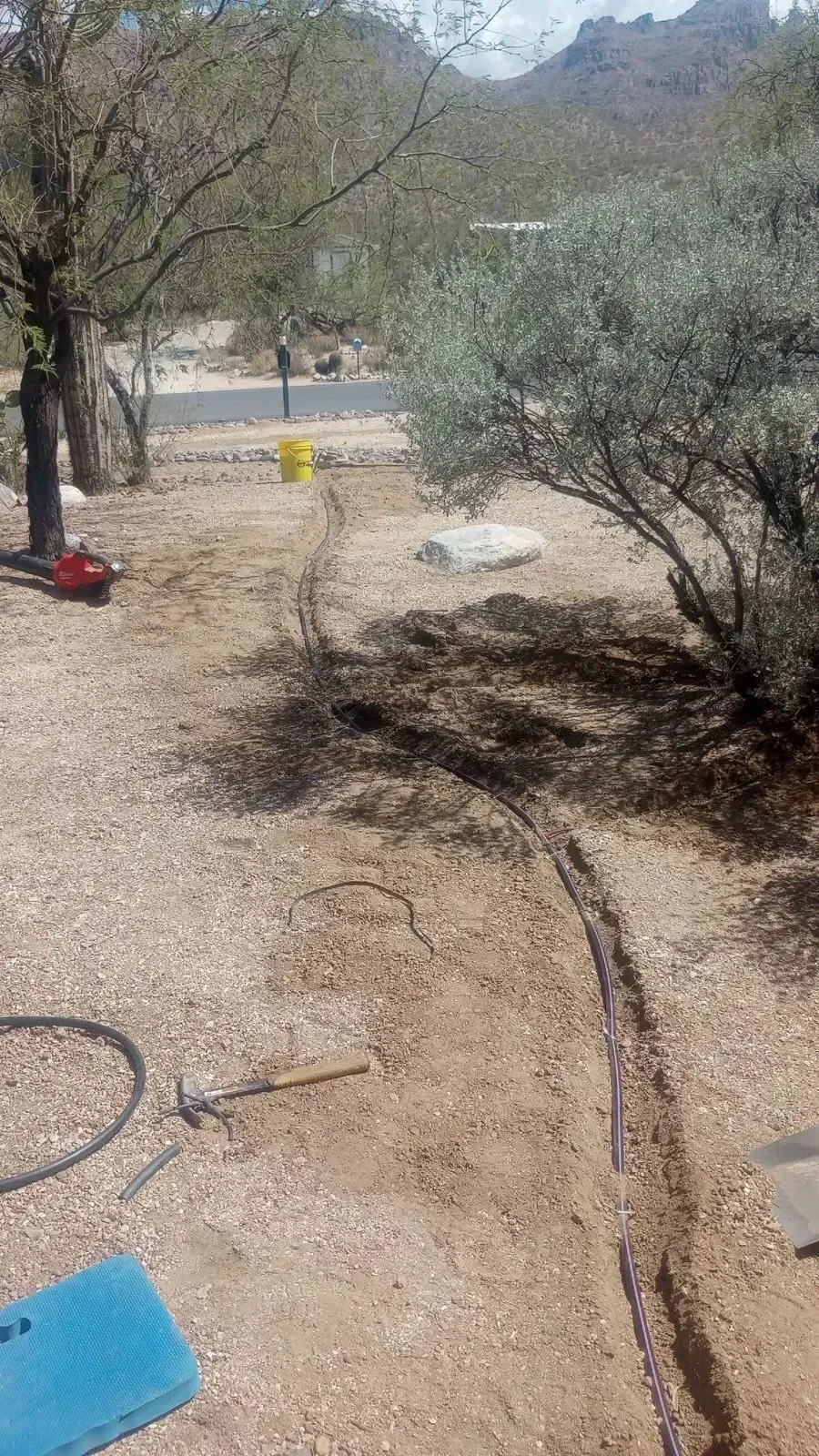 Trench in dirt with tools, trees, and distant road. Sunny day.