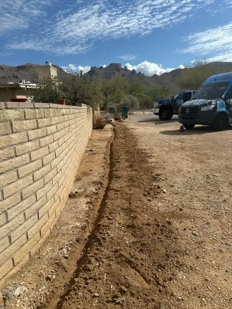 Trench dug in dirt next to a brick wall. Vans and a mountain range in the background under a blue sky.