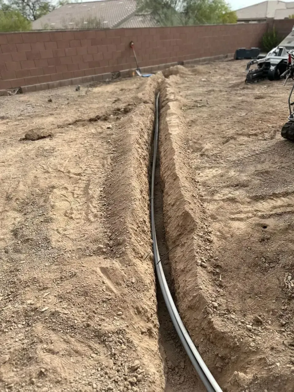 Trench dug in dirt, containing a gray pipe, possibly for utilities. Brown walls and a house are visible in the background.