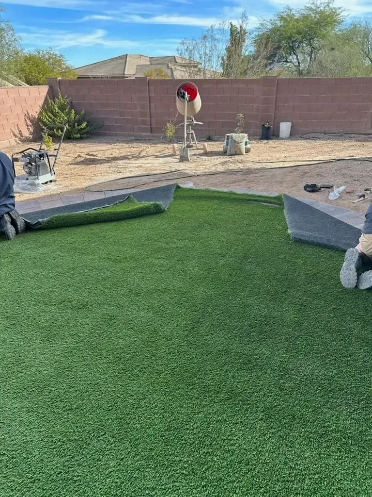 Installing artificial turf in a backyard. Green turf rolls are laid out. A red and white object stands in the center.
