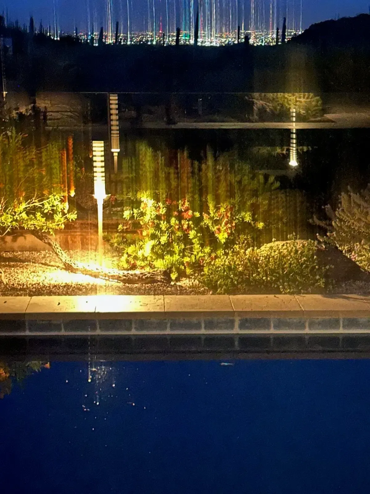 Night scene reflecting in water: illuminated landscaping, city lights in background, dark blue sky.