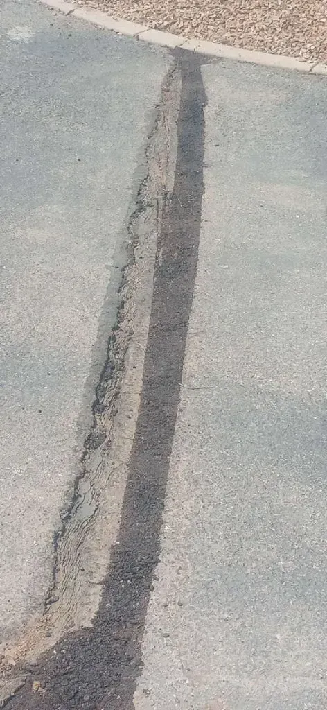 A long crack in asphalt pavement, partially shaded, with a gravel edge at top.