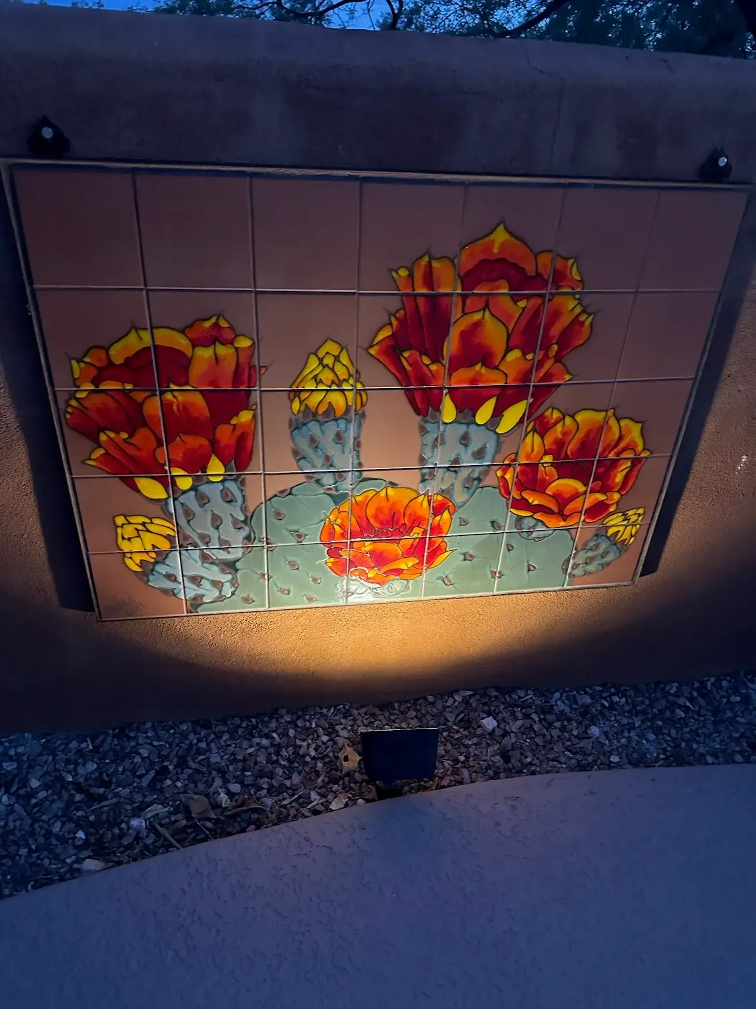 Tile mural of blooming cacti with orange flowers, lit by a spotlight, mounted on a wall.