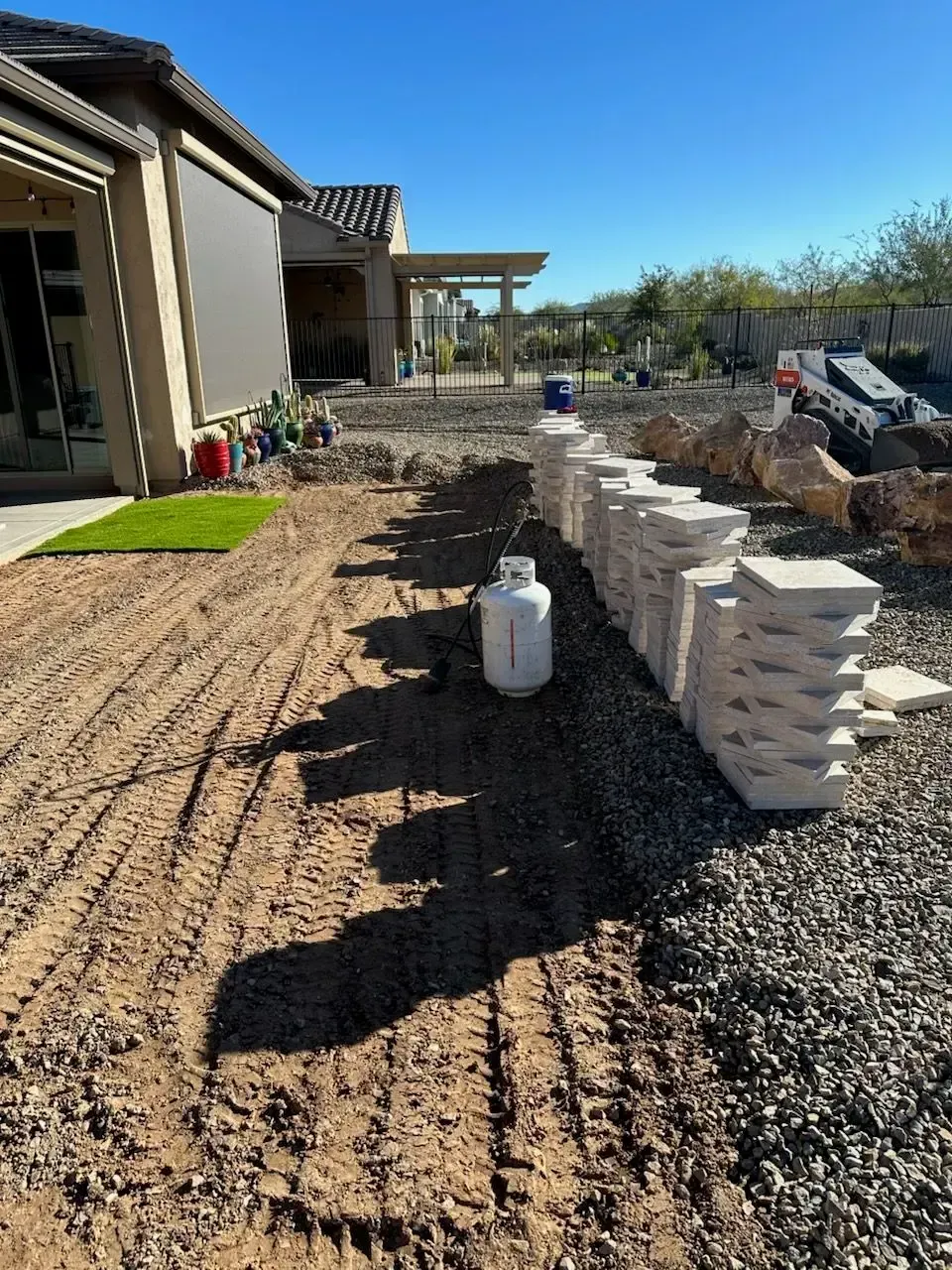 Construction site with stacked pavers, propane tank, and dirt path next to a house with outdoor living space.