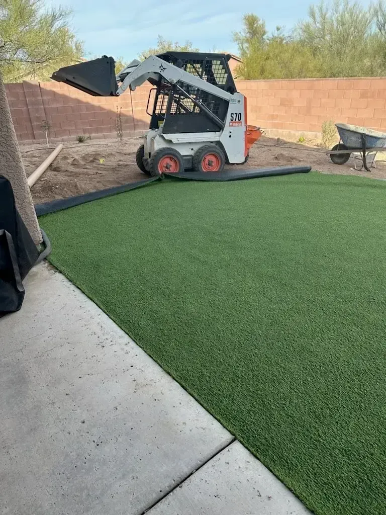 Bobcat moving soil next to new artificial turf; wheelbarrow and wall in the background.