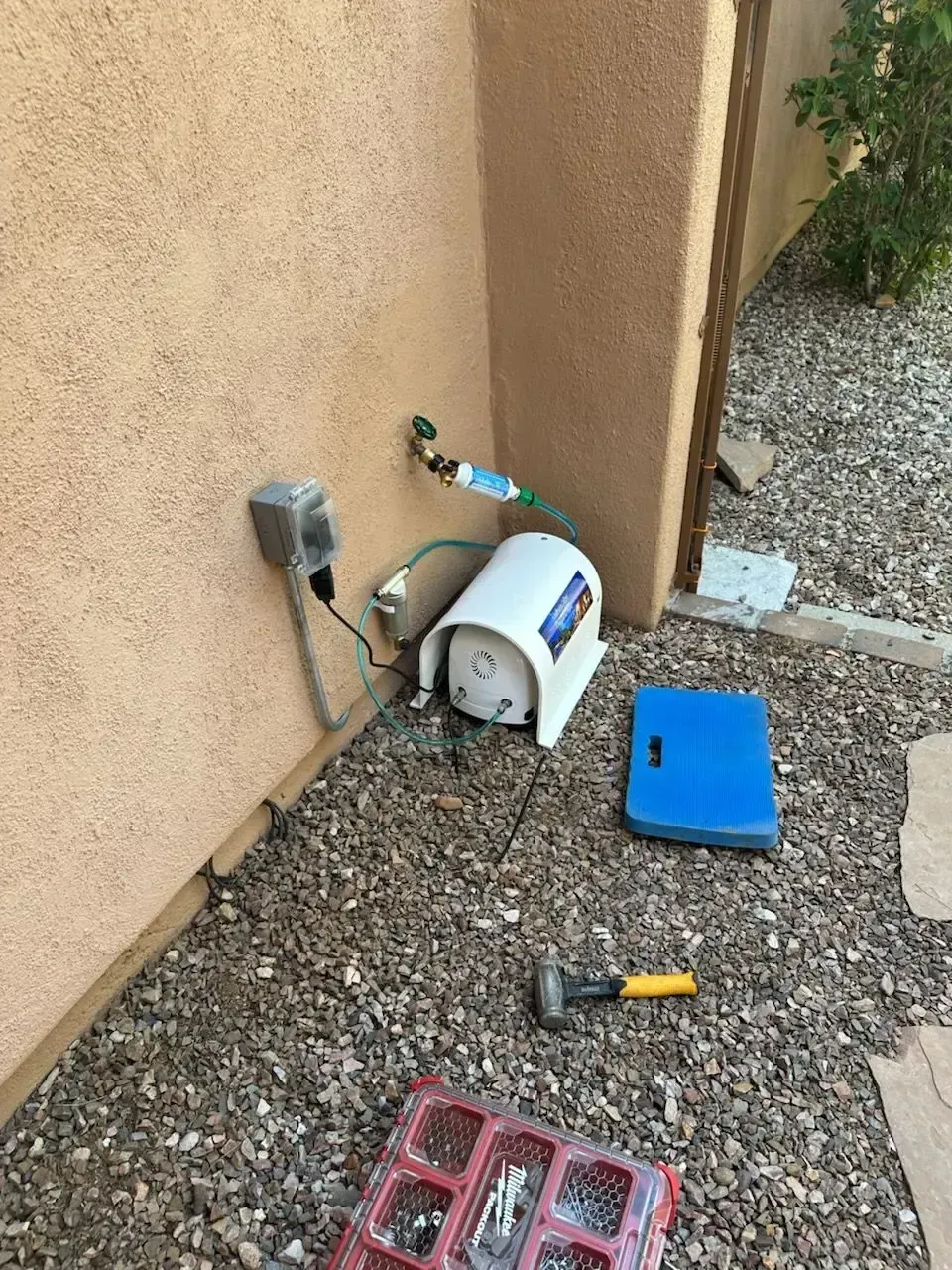 Outdoor water heater next to stucco wall, electrical outlet, and blue box on gravel ground.