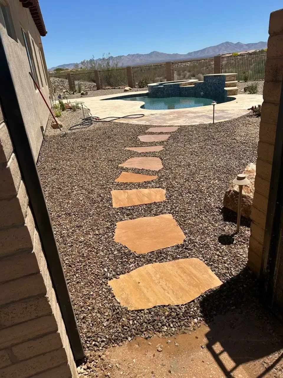 Flagstone pathway through gravel leading to a pool in a backyard. Blue sky and desert background.