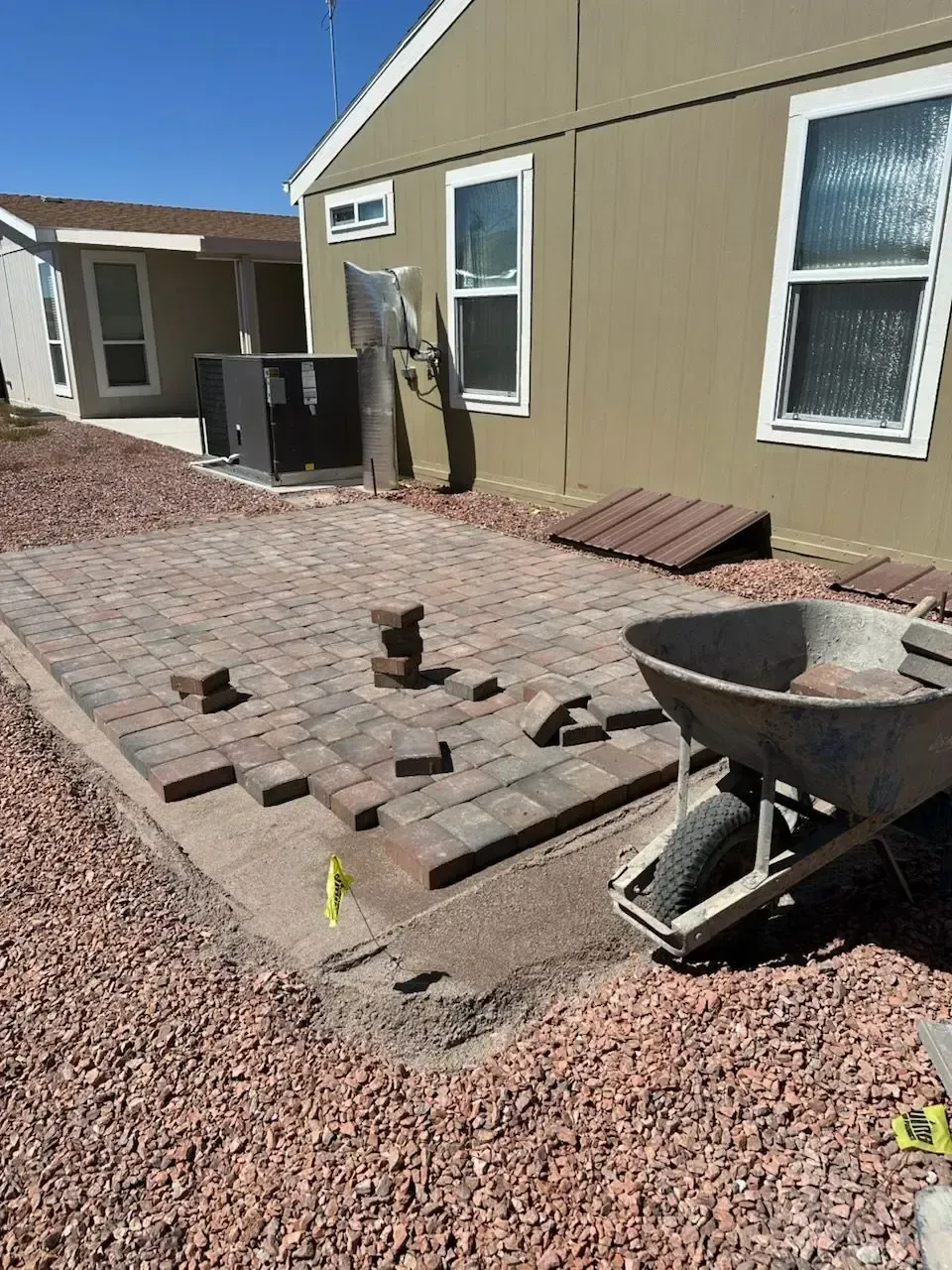Brick patio under construction next to a tan building with a wheelbarrow filled with sand and gravel.