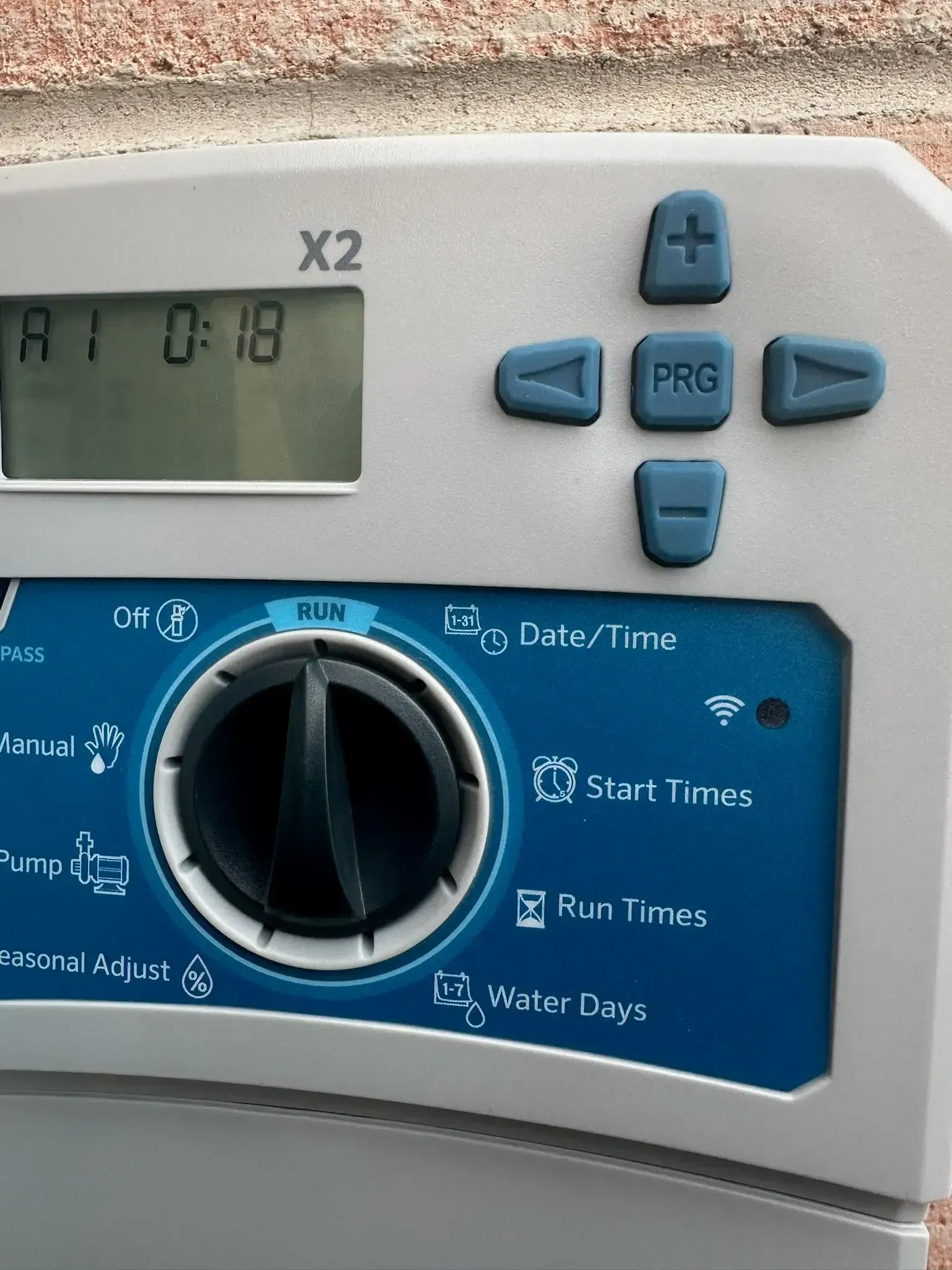 A digital sprinkler system control panel with a dial, buttons, and LCD screen against a brick wall.