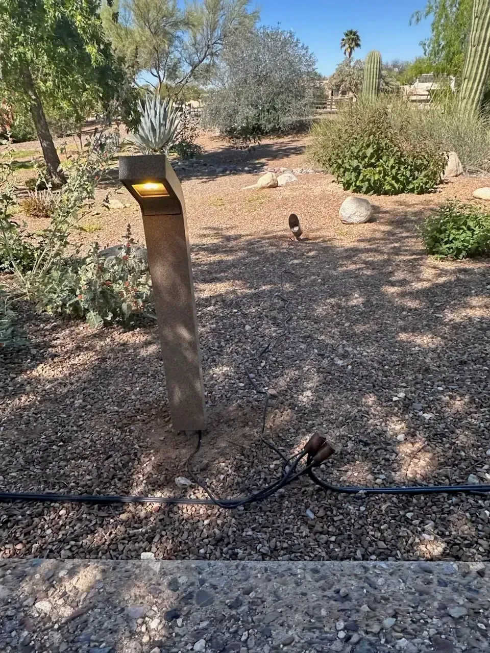 Pathway with a lit concrete light post and landscape plants, mostly brown gravel.