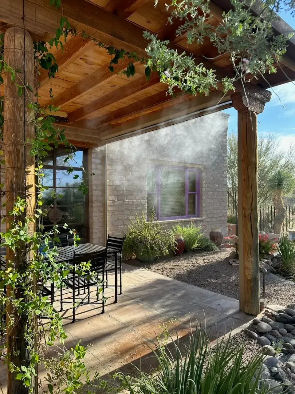 Outdoor patio with misting system, wooden beams, table, chairs, and plants; brick building in the background.