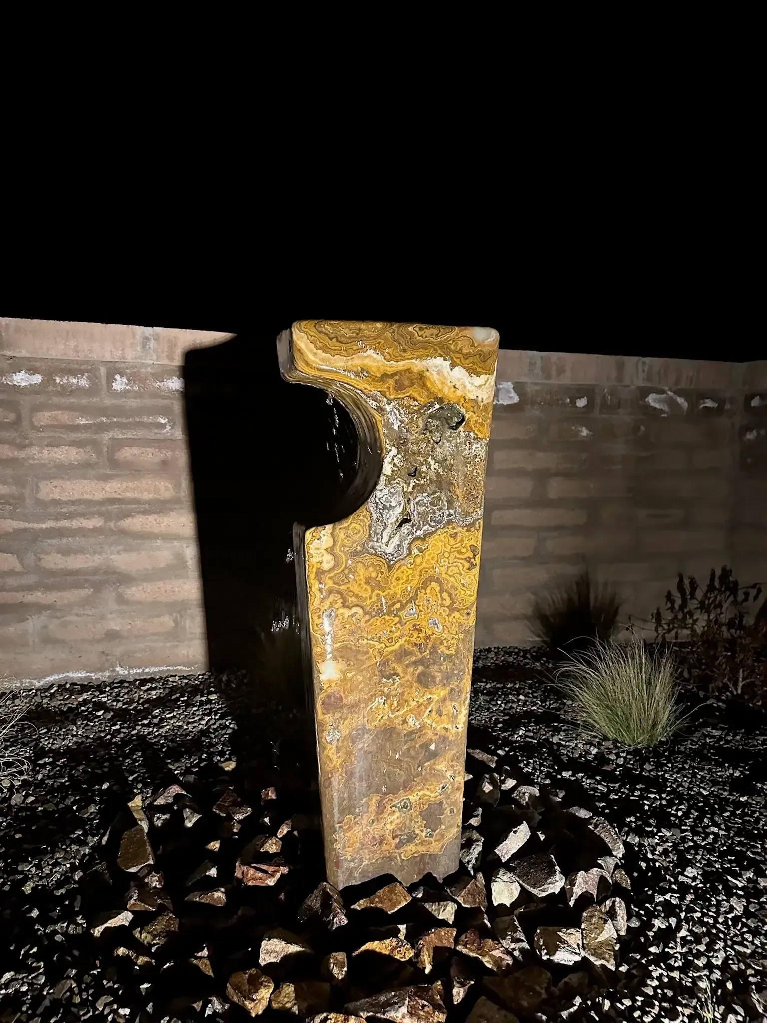 Tall, textured stone water fountain against a brick wall, surrounded by black rocks; at night.