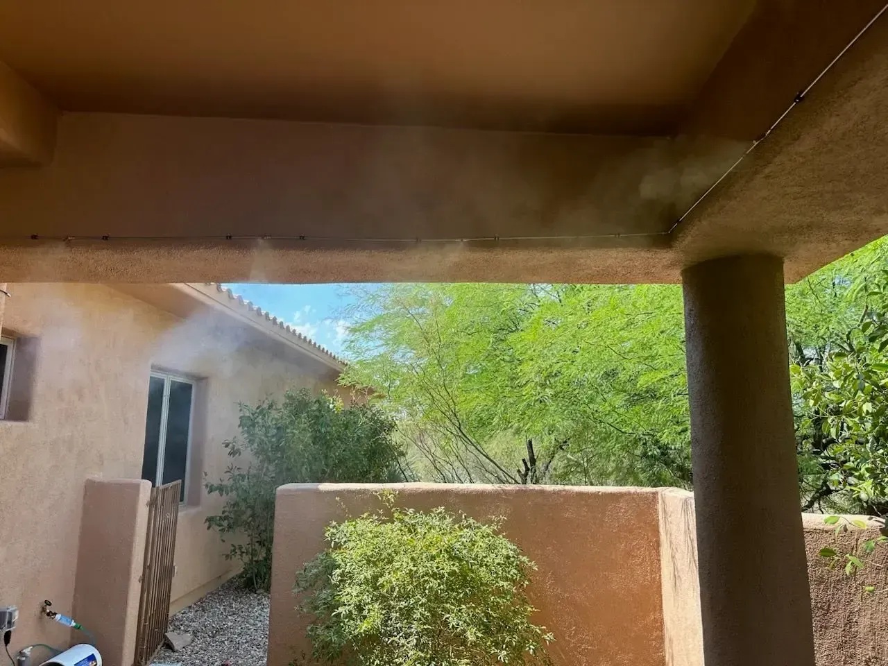 Patio with misting system, tan stucco walls, green bushes, and blue sky visible.