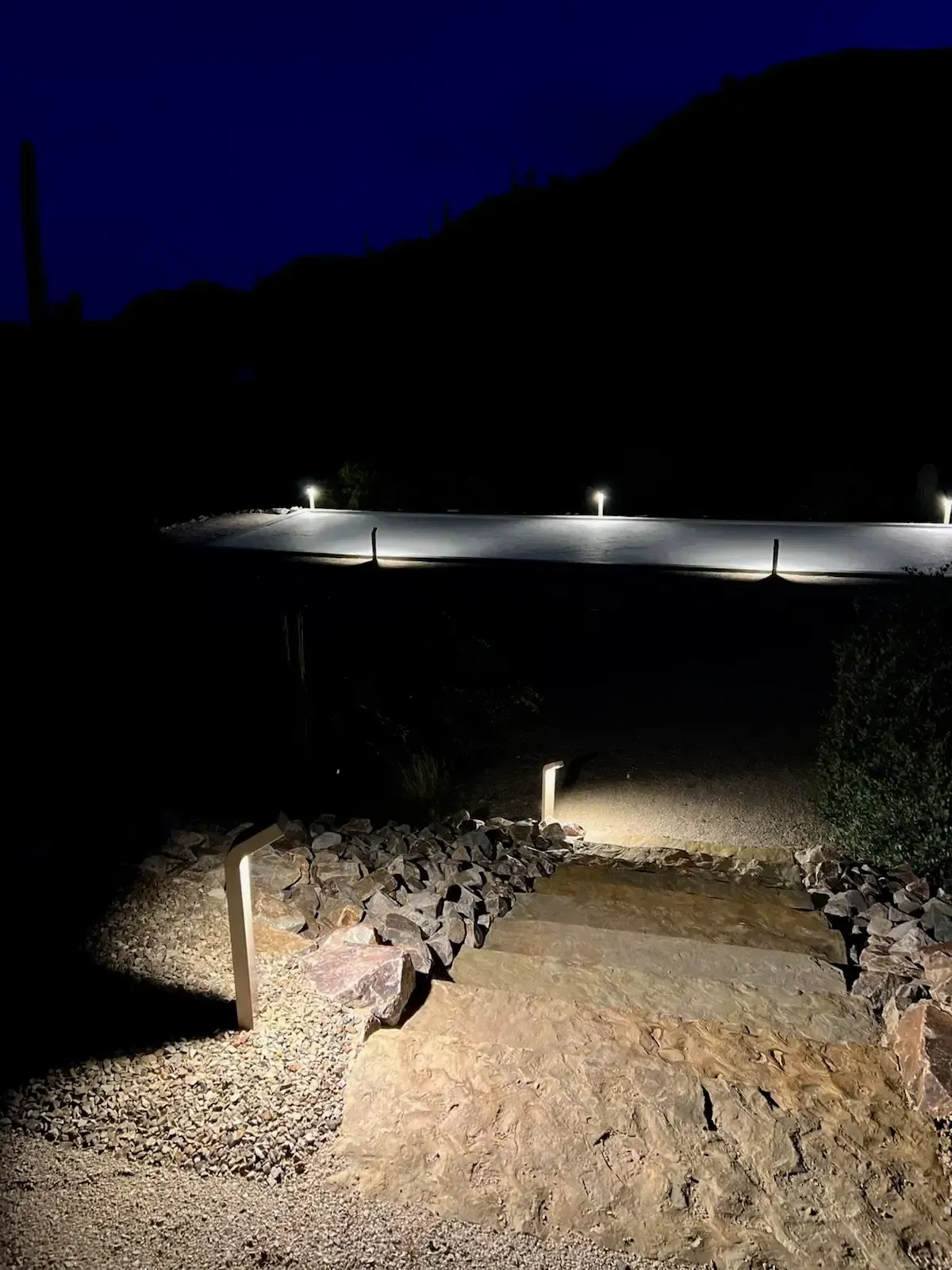 Gravel pathway lit by low lights at night, leading to more lights on a far path near a dark mountain.