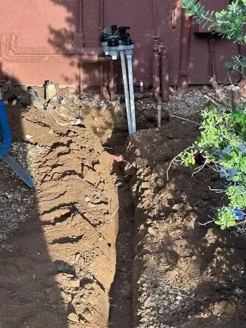 Trench dug in the dirt leads to irrigation system valves against a brown building.