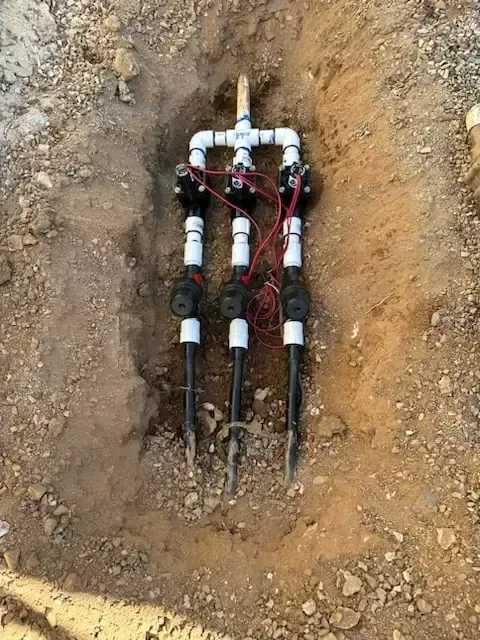 Three sprinkler valves connected with PVC piping in a brown dirt trench.