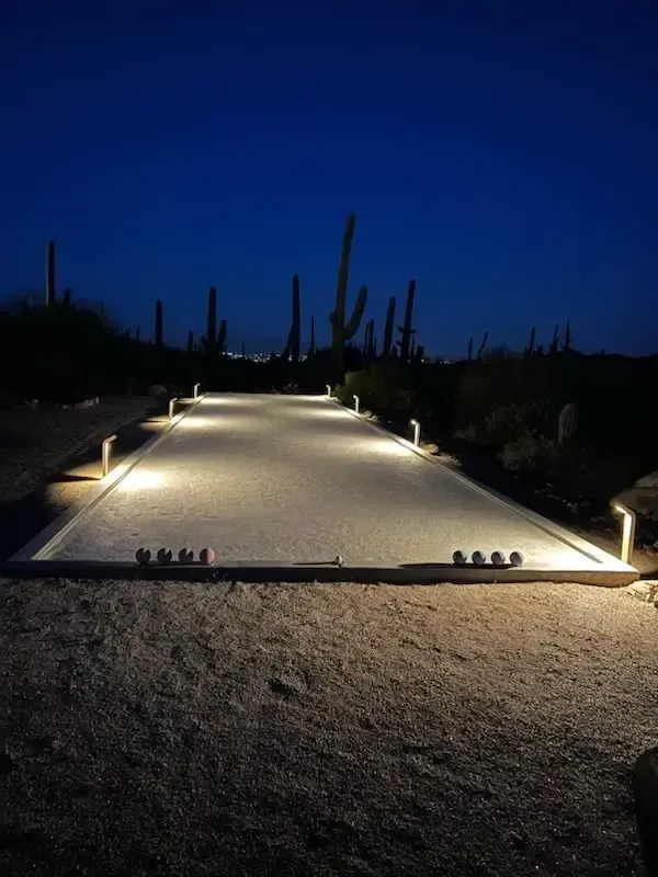 Nighttime view of a bocce ball court lit by overhead lights, with balls on the gravel surface and cacti in the background.