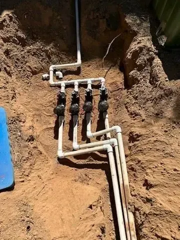 PVC irrigation system with four black valves in a dug-out area, against a sandy backdrop.