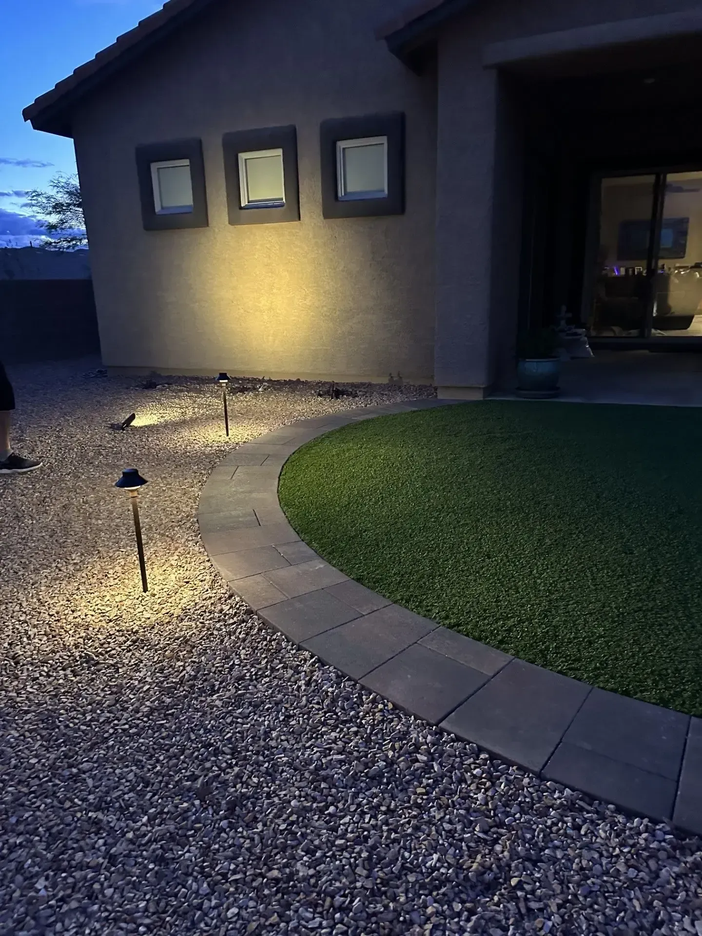 Outdoor scene at dusk with path lights illuminating a gravel bed, a curved walkway, and a green lawn.