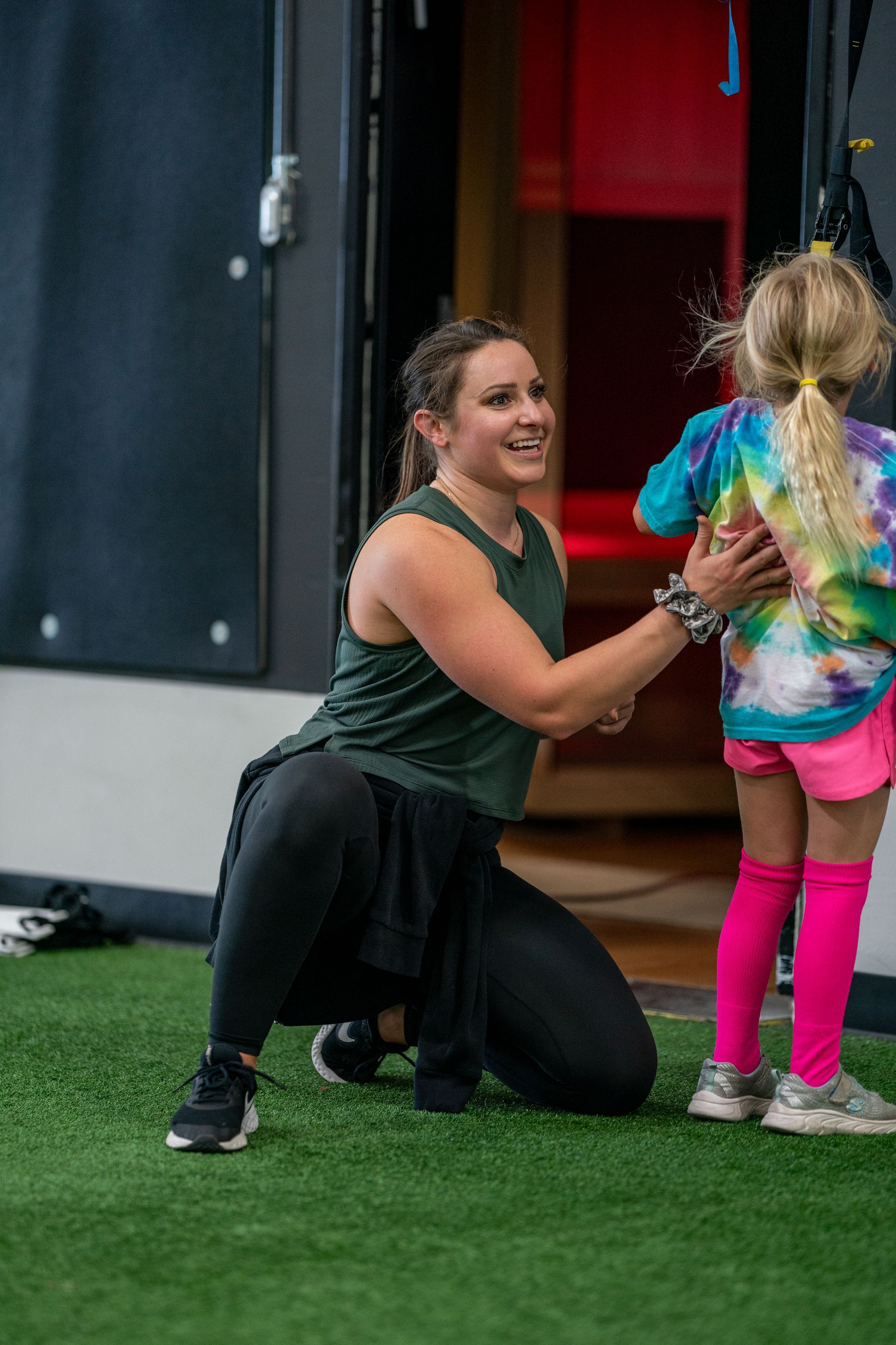 Woman kneeling and smiling as she helps a child in bright tie-dye clothes near a doorway on turf