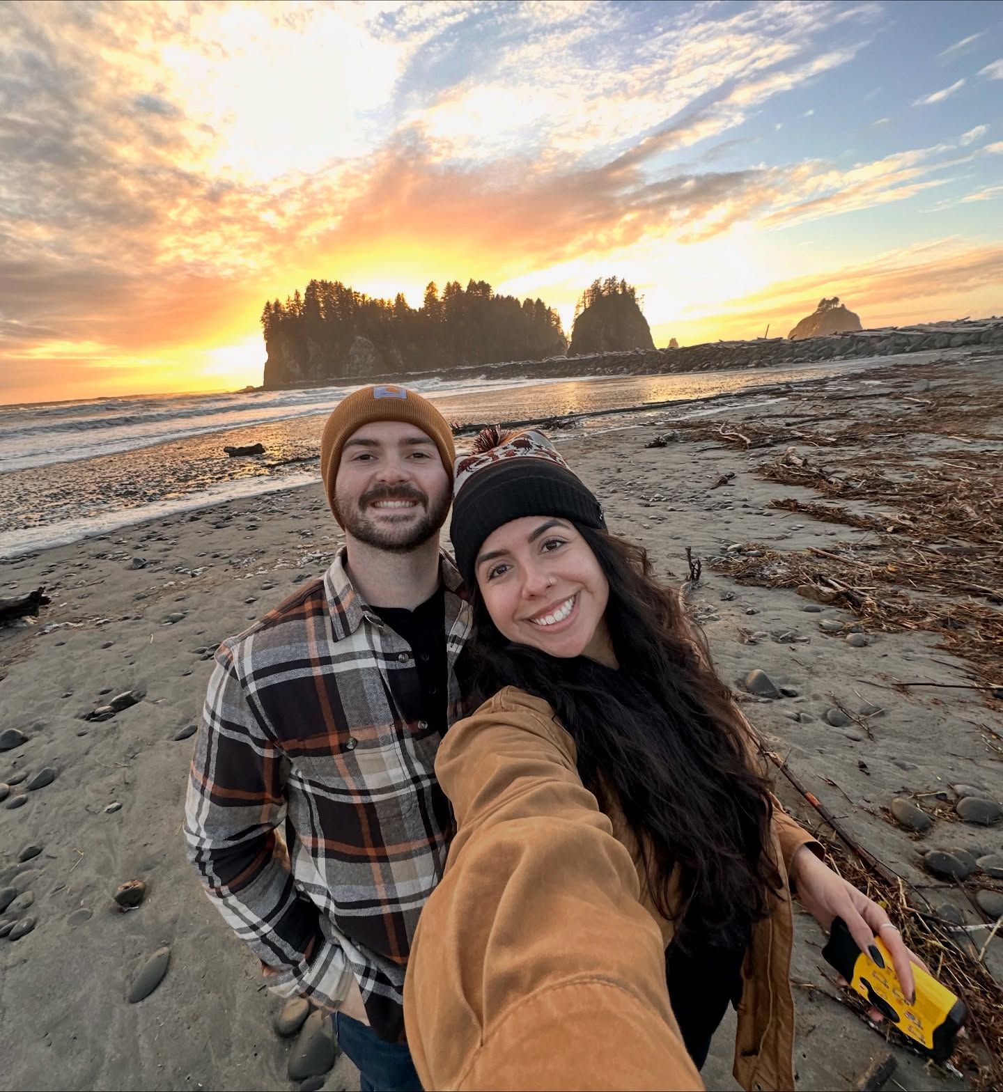 Selfie of two smiling people on a beach at sunset with sea stacks and waves in the background