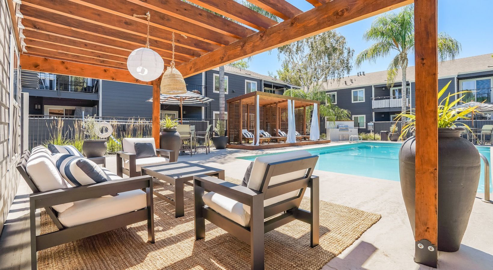 Outdoor pool area with seating under a wooden pergola, pool in background, and residential buildings visible.